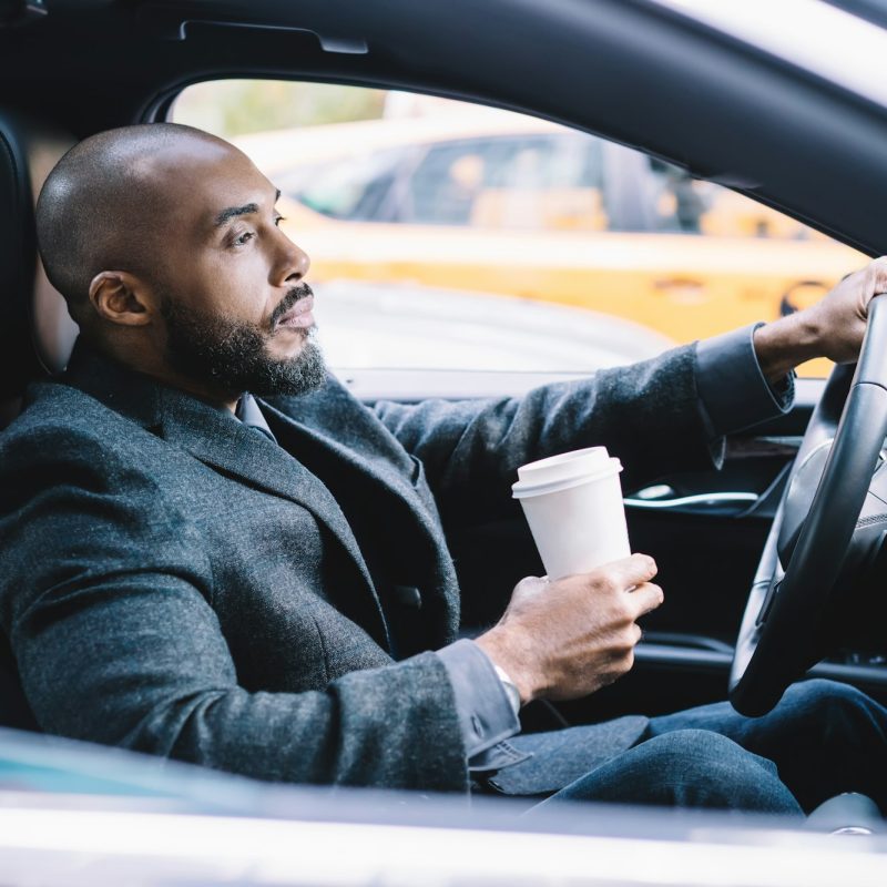 Calm pensive ethnic businessman with coffee to go driving car in city