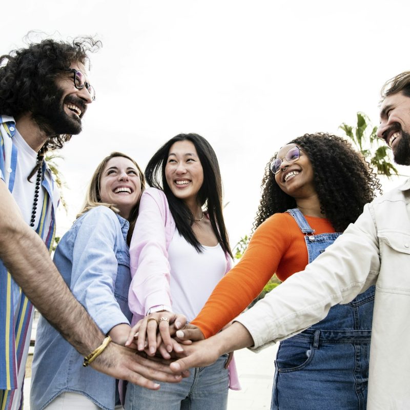 Diverse group of cheerful millennial friends standing together stacking hands in the street
